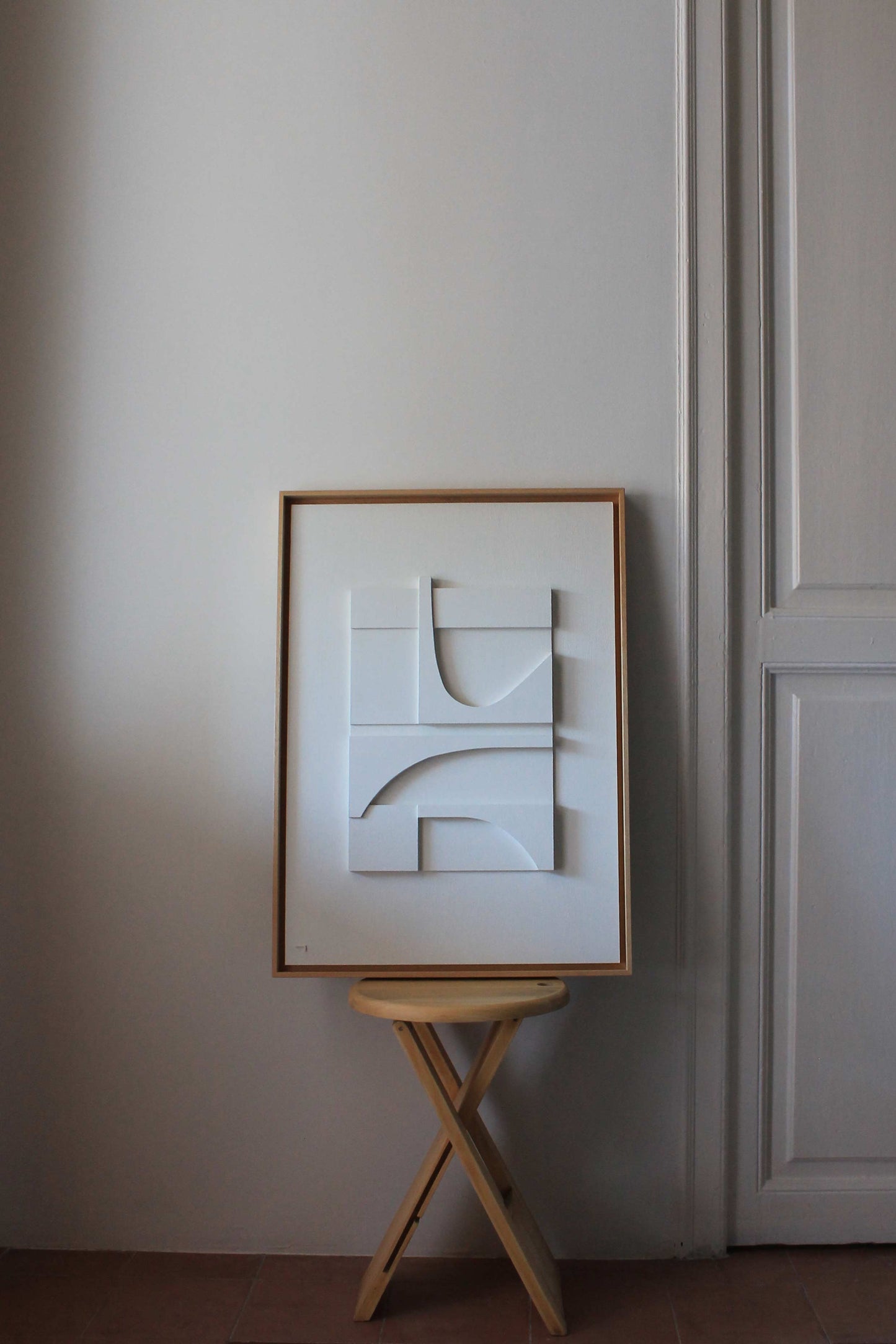 Cuadro blanco junto a una ventana, se proyectan sombras laterales entre las capas del relieve. La obra está enmarcada en madera natural. Aparece apoyada sobre un taburete junto a una puerta antigua con molduras.